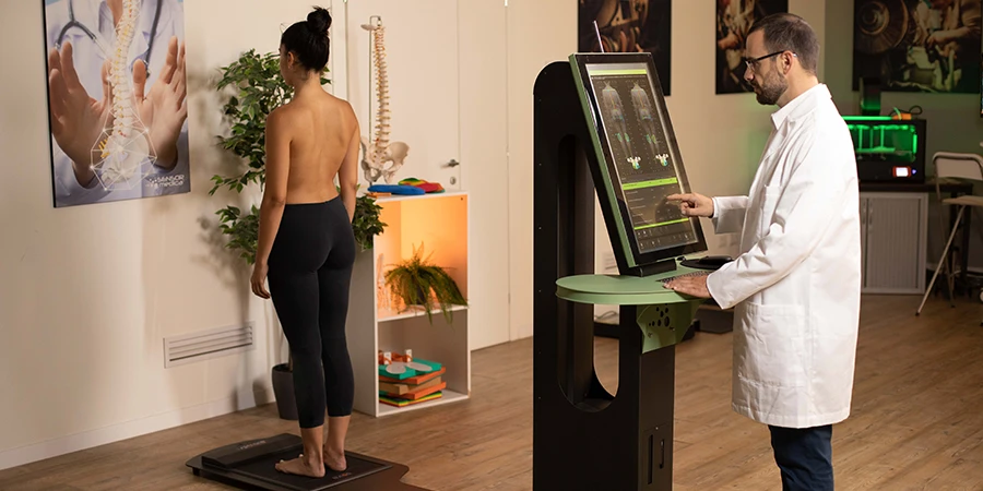 spine 3D dans un cabinet, quelqu'un en blouse touche l'écran et un patient torse nu de dos est sur la plateforme